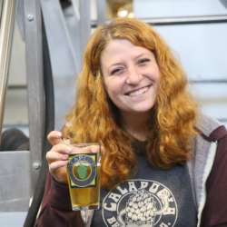 Portrait of Dani Raiche holding a beer she brewed and smiling into the camera at her place of work, Calapooia Brewing.