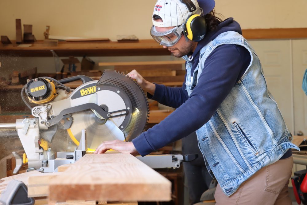Male student in a woodshop using a chop saw to cut a plank of wood.