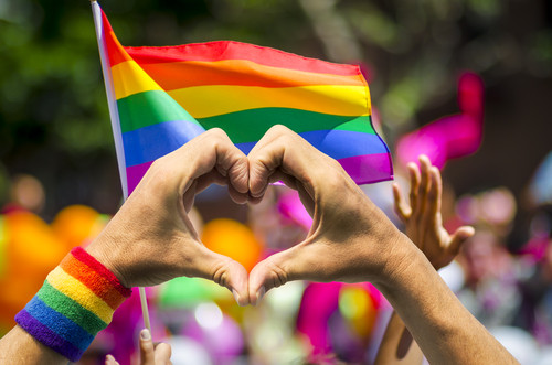 hands forming a heart shape with LGBTQI+ flag in background