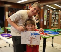 older and younger brother hold up their gyotaku print making project as they smile to the camera"
